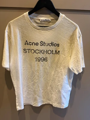 Vit Acne Studios t-shirt - Vit t-shirt från Acne Studios med tryck 'Acne Studios STOCKHOLM 1996' på bröstet. Klassisk passform och rund halsringning. Perfekt basplagg för en clean och stilren look.