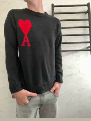 Svart stickad tröja från Ami Paris  - Svart stickad tröja från Ami Paris  med röd hjärtdetalj och bokstaven A på bröstet. Tröjan har rund halsringning, ribbade muddar och är perfekt för dig som gillar stilrena plagg med en twist.