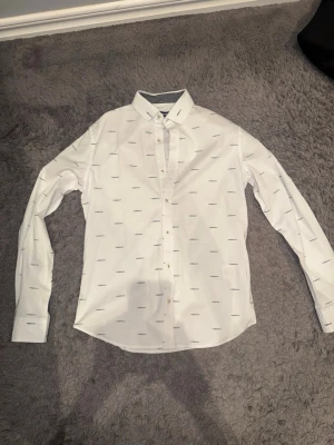 Vit skjorta från DKNY med logotypmönster - Säljer en vit skjorta från DKNY med diskret logotypmönster över hela plagget. Skjortan har klassisk krage, långa ärmar och knäppning framtill. Materialet är mjukt och känns som bomull, perfekt för en clean och stilren look.