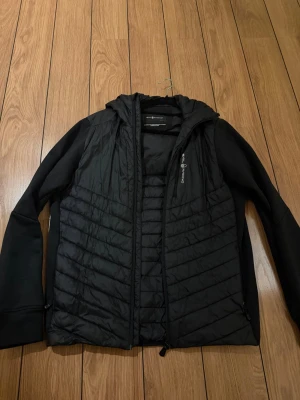 Jackor  - This is a black Sail Racing jacket in size Medium. The jacket features a quilted front and hood, with softshell sleeves and sides. It has a zippered chest pocket and is designed by Sail Racing International in Sweden, assembled in China.