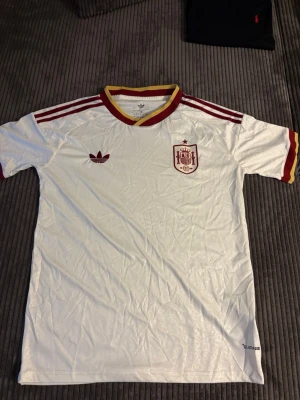 Vit Spanien tröja Adidas M - Säljer en vit Spanien fotbollströja från Adidas med röda och gula detaljer. Tröjan har korta ärmar, röd Adidas-logga och Spaniens landslagsemblem på bröstet. Klassiska tre ränder på axlarna och ribbad krage i rött och gult. Perfekt för match eller träning.