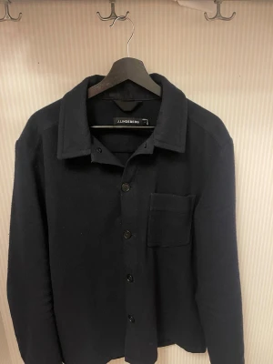 Mörkblå overshirt från J.Lindeberg - Stilren mörkblå overshirt från J.Lindeberg med klassisk krage, knappstängning och en bröstficka. Tillverkad i ett mjukt, tjockare tyg som passar perfekt som lager. Enkel och clean design som funkar till många olika looks.