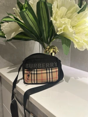 Burberry axelväska med rutigt mönster - Snygg axelväska från Burberry med klassiskt beige, svart och rött rutmönster på framsidan. Väskan har svart kant och justerbar axelrem. Stor logga på ovansidan och dragkedja med rödvit detalj. Perfekt för dig som gillar streetstyle och designeraccessoarer. Helt ny 