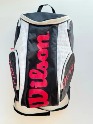 Wilson svart och vit ryggsäck - Snygg ryggsäck från Wilson i svart och vitt med stora röda logotyper. Ryggsäcken har flera fack med dragkedja och vadderade axelremmar för extra komfort. Perfekt för sport eller skola, tillverkad i slitstarkt syntetmaterial.