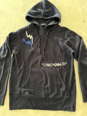 Mörkblå hoodie från Monalto med tryck - Mörkblå hoodie från Monalto med huva och dragsnören. Framsidan har ett vitt och blått blixtmönster samt texten 'L'ART MONALTO' tryckt i vitt. Praktisk magficka framtill. Perfekt för en chill och trendig look.
