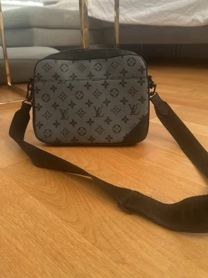 Grå axelväska med monogram sällsynt modell  - Snygg grå axelväska med svart monogrammönster och justerbar bred axelrem. Väskan har en rektangulär form och dragkedja upptill. Perfekt för dig som vill ha en stilren och trendig accessoar till vardagslooken.