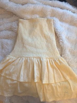Gul axelbandslös volangklänning - Söt gul axelbandslös klänning med smockad överdel och dubbla volanglager i kjolen. Klänningen har fina broderade detaljer i tyget och är perfekt för varma dagar. Tillverkad i bomull för en luftig och bekväm känsla. Använd 2 gånger🥰