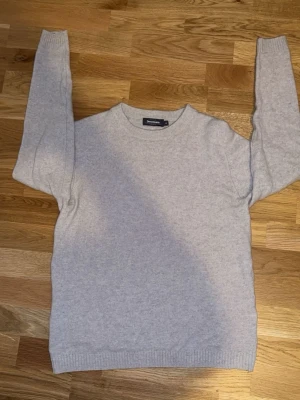 Ljusgrå stickad tröja Dressmann - En stilren ljusgrå stickad tröja från Dressmann i 100% Bomhus och är i en klassisk modell. Tröjan har rund halsringning och ribbade muddar vid ärmslut och nederkant. Perfekt att matcha med jeans eller chinos för en clean look.