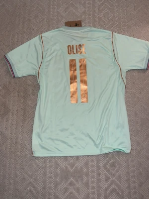 Olise.11 tröja frankrikes bortaställ - Snygg ljusblå fotbollströja från Nike med Frankrikes landslagsmärke och guldiga detaljer. Tröjan har korta ärmar, guldiga ränder och nummer 11 samt namnet 'OLISE' på ryggen. Perfekt för dig som älskar fotboll och vill sticka ut på planen.