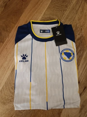 Bosnien & Hercegovina matchtröja (Kelme) – Helt ny med lappar! - Tröjan har en riktigt snygg design i vitt, blått och gult med finstämda ränder och förbundsmärket på bröstet. 🔥 Passa på att köpa nu i god tid innan landskampen mellan Sverige och Bosnien på Strawberry Arena! Perfekt att ha på sig på läktaren för att visa ditt stöd.
