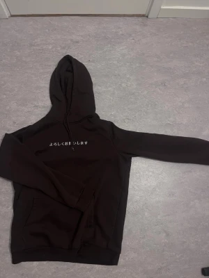 Svart hoodie med japansk text - Säljer en svart hoodie med huva och känguruficka framtill. På bröstet finns vit japansk text tryckt. Hoodien har dragsko i huvan och är gjord i mjukt material, perfekt för chill dagar.