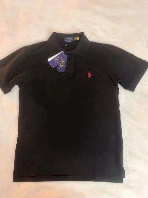 Svart pikétröja från Polo Ralph Lauren - Klassisk svart pikétröja från Polo Ralph Lauren med röd broderad logga på bröstet. Tröjan har korta ärmar, krage och knappar vid halsen. Tillverkad i bomull för en bekväm och stilren look.