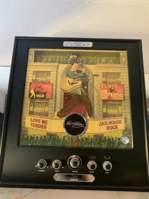 Elvis Presley radio tavla retro - Cool radio inbyggd i en tavla med Elvis Presley-motiv. Tavlan har svart ram och färgglad illustration av Elvis med gitarr, samt detaljer som 'Love Me Tender' och 'Jailhouse Rock'. Utrustad med silverfärgade rattar för volym, ton, tuning och band. Perfekt för musik- och retrostyle collectible 