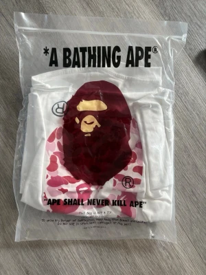 Vit BAPE t-shirt med rosa camo print - Säljer en vit t-shirt från A Bathing Ape med ikoniskt rosa camo-apansikte och en liten apa ovanpå trycket. T-shirten har rund hals och korta ärmar. Perfekt för dig som gillar streetwear och vill sticka ut med en unik design.