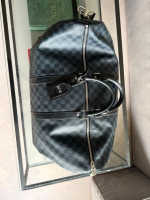 Louis Vuitton Keepall 55 - Snygg svart weekendbag med klassiskt rutmönster och detaljer i svart läder. Väskan har dubbla handtag, silverfärgade dragkedjor och en bagagetagg. Perfekt storlek för resor och enkel att bära med sig. Service kvitto ifrån Louis Vuitton Stockholm finns. 