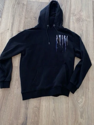 Svart hoodie från Amiri (L) - Säljer en svart hoodie från Amiri med färgstänk och logga både fram och bak. Hoodien har huva med dragsko, känguruficka och ribbade muddar. Materialet ser ut att vara mjukt och bekvämt, perfekt för en avslappnad streetwear-look.