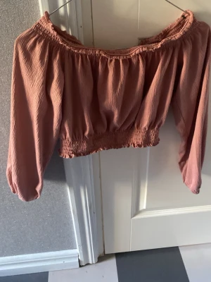 Rosa croppad offshoulder topp - Trendig rosa croppad topp med offshoulder-design och långa ärmar. Toppen har resår vid axlar och midja för en snygg passform och är gjord i ett lätt, skrynkligt tyg som ger en avslappnad känsla.