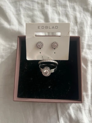 Smyckesset från Edblad med stenar -  HELT NYAAAA !!Snyggt smyckesset från Edblad med matchande örhängen och två ringar i silverfärgad metall. Alla delar har gnistrande klara stenar i cirkelformade mönster som ger en elegant look. Perfekt för dig som gillar stilrena accessoarer med lite extra bling. Värda 700-900kr