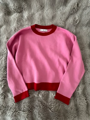 Rosa tröja med röda detaljer - Säljer en snygg rosa tröja med kontrasterande röda muddar, nederkant och halsringning. Tröjan har en rak passform och långa ärmar. Perfekt för dig som gillar färg och vill sticka ut lite extra.
