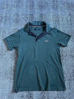Grön piké från Hugo Boss - Snygg grön pikétröja från Hugo Boss Selection med korta ärmar och klassisk krage. Tröjan har diskret Boss-logga på bröstet och kontrasttyg med små prickar på insidan av kragen. Perfekt för en stilren och avslappnad look.