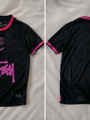 Svart t-shirt med rosa detaljer - Snygg svart t-shirt med korta ärmar och rosa detaljer vid krage och ärmslut. T-shirten har ett diskret mönster i tyget och en stor rosa logga framtill. Perfekt för dig som vill sticka ut med en unik stil.