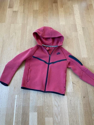 Röd hoodie med dragkedja från Nike - Snygg röd hoodie från Nike med svart logga på bröstet och svarta detaljer längs dragkedjan och ärmarna. Hoodien har hel dragkedja, två sidofickor och en extra ficka på ena ärmen. Tillverkad i mjukt och stretchigt material. Perfekt för en sportig look.