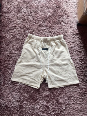 Beige shorts från Essentials - Säljer ett par beige shorts från Essentials med elastisk midja och diskret logga broderad framtill. Shortsen har en relaxed fit och är tillverkade i mjuk bomull, perfekta för chill dagar. Snygg minimalistisk design med vita detaljer.