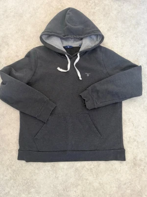 Mörkgrå hoodie från GANT 160 - Mörkgrå hoodie från GANT med vit dragsko och broderad logga på bröstet. Hoodien har en klassisk känguruficka framtill och mjukt grått innerfoder i huvan. Perfekt för en avslappnad och stilren look Passar dig som är runt 155-165 och priset går o sänka  .