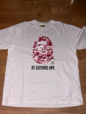 Vit BAPE t-shirt med rosa camo print - Säljer en vit t-shirt från BAPE med det klassiska apehuvudet i rosa och röd camo på bröstet samt texten 'BY BATHING APE' under. T-shirten har rund hals och korta ärmar. Perfekt för dig som gillar streetwear och vill sticka ut.
