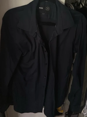 Svart skjorta/kavaj från BOSS - Stilren svart skjorta från BOSS med klassisk krage och knappar framtill. Skjortan har lång ärm och är tillverkad i ett mjukt material som känns bekvämt mot huden. Perfekt för dig som vill ha en enkel men snygg look.