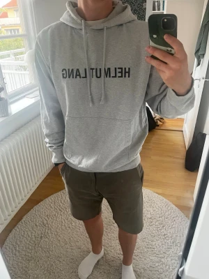 Grå hoodie från Helmut Lang - Snygg grå hoodie från Helmut Lang med svart logotyp framtill. Hoodien har klassisk känguruficka, justerbar huva med snörning och ribbade muddar. Perfekt för en chill och stilren look. Ny hoodie kostar mellan 4000-7000 kronor