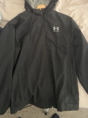 Svart regnjacka från Under Armour - Snygg svart regnjacka från Under Armour med huva och dragkedja framtill. Jackan har meshfoder på insidan för extra ventilation och två fickor med dragkedja. Perfekt för regniga dagar och sportiga aktiviteter.