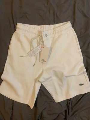 Vita shorts med snörning från Lacoste - Snygga vita shorts från Lacoste med klassisk krokodillogo på benet. Shortsen har elastisk midja med snörning och sidofickor. Perfekta för sommaren och har en clean, sportig vibe.