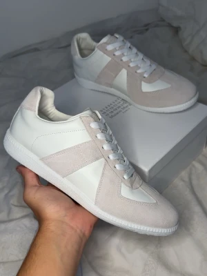 Maison margiela  - Snygga sneakers i vitt och beige med ovandel i både mocka och läder. Klassisk design med vita snören och vit sula. Perfekta för en clean och stilren look. Låg modell som passar till det mesta och ger en fräsch vibe.