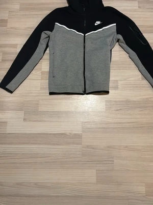Nike grå och svart hoodie med dragkedja - Snygg hoodie från Nike i grått och svart med vit logga på bröstet. Tröjan har hel dragkedja framtill, huva och en vit vågig detalj över bröstet. Perfekt för en sportig och avslappnad stil.
