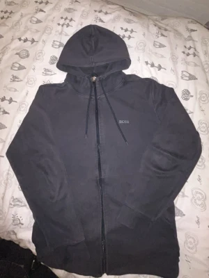 Svart hoodie med dragkedja från BOSS - Säljer en svart hoodie från BOSS med dragkedja framtill och justerbar huva med snörning. Hoodien har diskret BOSS-logga på bröstet och är långärmad. Perfekt för en avslappnad och stilren look.