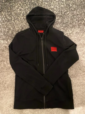 Svart hoodie med dragkedja från Hugo - Äkta hoodie från Hugo med röd logopatch på bröstet. Modellen har huva med dragsko, hel dragkedja framtill och långa ärmar. Tillverkad i mjukt material som känns skönt mot huden. Perfekt för en avslappnad och stilren look.