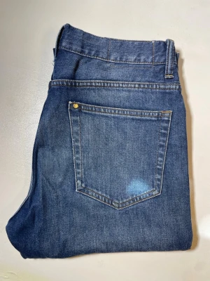Blå raka jeans i klassisk modell - Snygga blå jeans med rak passform och fem fickor. Jeansen har en klassisk midja, bälteshällor och subtila slitningar framtill för en avslappnad look. Tillverkade i denim med kontrastsömmar och metallknappar.