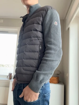 Moncler grå cardigan - Moncler cardigan i mörkgrått med quiltad framsida och mjuka stickade ärmar och rygg. Hög krage, dragkedja framtill och två sidofickor med dragkedja. Klassisk Moncler-logga på vänster ärm för en clean och exklusiv vibe.