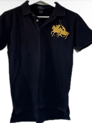 Mörkblå Ralph Lauren pike  - Snygg mörkblå pikétröja från Ralph Lauren med korta ärmar och stor gul broderad logga på bröstet. Klassisk krage och knappar. Perfekt för en avslappnad men stilren look.