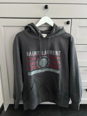 Grå Saint Laurent hoodie med tryck - Säljer en grå hoodie från Saint Laurent med huva och dragsko. Framsidan har ett stort tryck med texten 'SAINT LAURENT' och röda samt vita detaljer. Hoodien har en klassisk känguruficka och ribbade muddar. Perfekt för en avslappnad och trendig look.