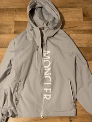 Ljusgrå vindjacka från Moncler - Snygg ljusgrå vindjacka från Moncler med stor vit logga längs dragkedjan. Jackan har huva, två framfickor med dragkedja och justerbar nederkant. Tillverkad i ett lätt, vindtätt material med slät insida. Perfekt för dig som vill ha en stilren och sportig look.