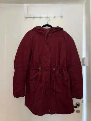 Vinröd parkas med huva och dragsko - Vinröd parkas med huva och dragsko i midjan. 