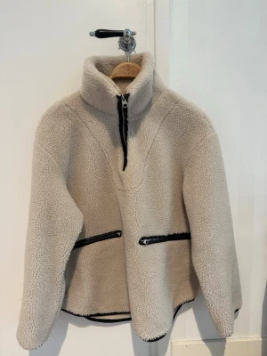 Beige teddyjacka  - Mysig beige teddyjacka från arket med hög krage och halv dragkedja framtill. Två snedställda fickor med svarta dragkedjor och svarta detaljer längs kanterna. Tillverkad i fluffigt teddy-material som håller dig varm och stylish.