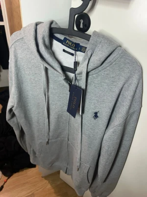 Grå hoodie från Polo Ralph Lauren - Snygg grå hoodie från Polo Ralph Lauren med dragkedja framtill och klassisk blå logga broderad på bröstet. Hoodien har huva med dragsko och två fickor framtill. Tillverkad i mjuk bomullsblandning, perfekt för en chill och stilren look.