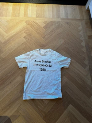 Acne Studios Stockholm - Vit t-shirt från Acne Studios med svart tryck på bröstet där det står 'Acne Studios STOCKHOLM 1996'. Klassisk passform och rund hals. Perfekt till jeans eller shorts för en clean och enkel look.