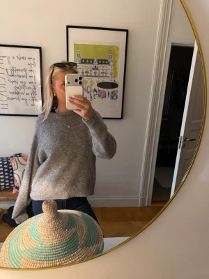 Grå stickad tröja med rund hals - Mysig grå stickad tröja med rund hals och långa ärmar. Tröjan har en avslappnad passform och ribbstickade muddar vid ärmslut och nederkant. Perfekt för kyliga dagar och enkel att matcha med jeans eller kjol.