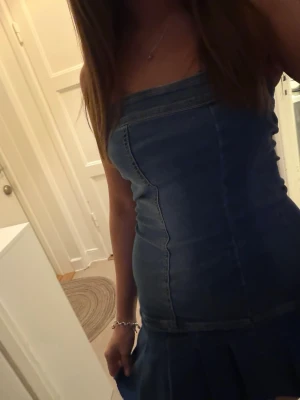 Jeansklänning axelbandslös kort - Säljer en snygg axelbandslös jeansklänning i blå denim. Klänningen är kort med figurnära passform och har en volangliknande nederdel med råa kanter. Perfekt för dig som vill ha en trendig och ungdomlig look.