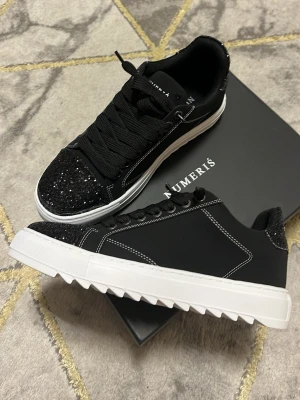 Numeris (svart glittriga) skor - Snygga svarta sneakers från Numeris med glittriga detaljer på tå och häl. Skorna har vita sicksacksulor, svarta skosnören och kontrastsömmar. Perfekta för dig som vill sticka ut med en unik och trendig look. Helt nya och oanvända!