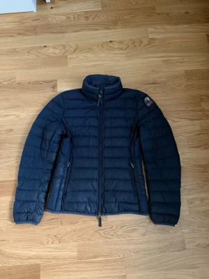 ÄKTA Mörkblå dunjacka från Parajumpers - Säljer en mörkblå dunjacka från Parajumpers i superlätt material. Jackan har hög krage, två sidofickor med dragkedja och quiltad design. Märkespatch på ärmen och snygga detaljer som orange ögla i nacken. Perfekt för kalla vinterdagar.
